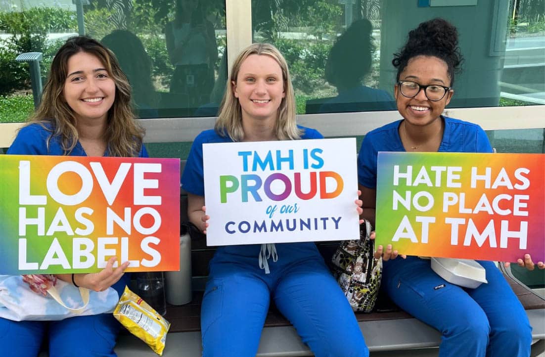 Three people in blue scrubs sit on a bench holding colorful signs. The signs read "LOVE HAS NO LABELS," "TMH IS PROUD of our COMMUNITY," and "HATE HAS NO PLACE AT TMH." They are smiling, and the background shows greenery through a window.