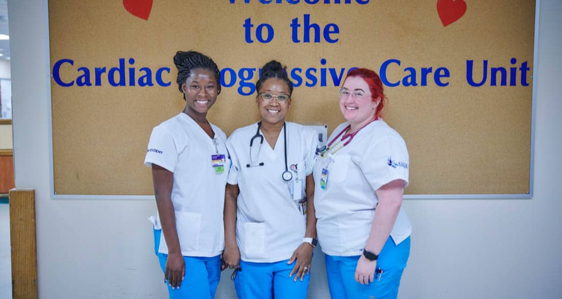 Three healthcare professionals wearing white uniforms and blue pants stand in front of a bulletin board that reads "Welcome to the Cardiac Progressive Care Unit." They are smiling and posing for the photo. The board is decorated with small red hearts.