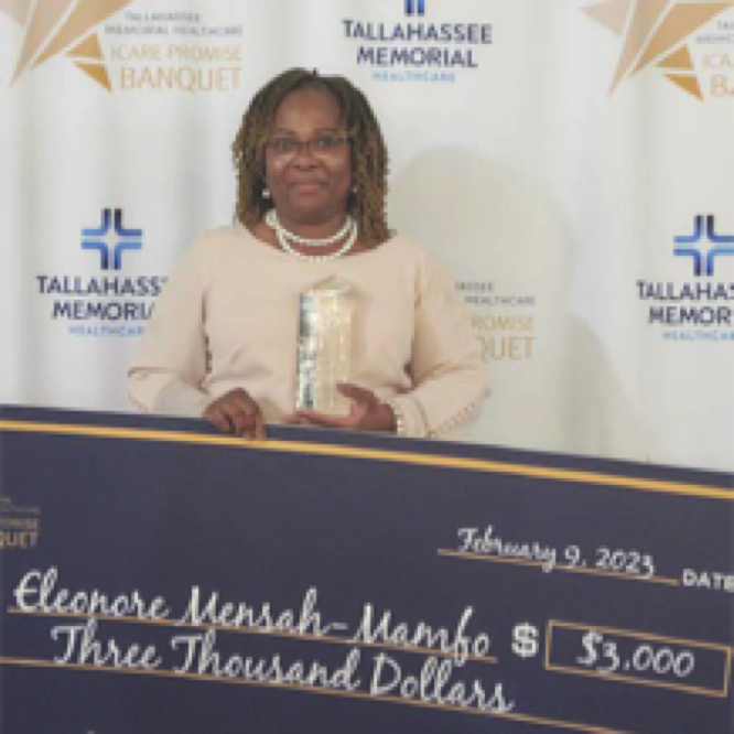 A woman stands holding an award and a large ceremonial check for $3,000 at the Tallahassee Memorial Healthcare 2023 ICARE Promise Banquet. The check is made out to Elcopore Mensah-Mamfo. The event backdrop features the organization's logo and event name.