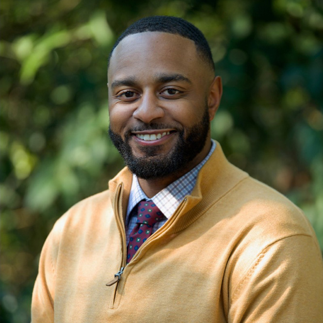 A man with a short beard and mustache is smiling at the camera. He is wearing a tan sweater over a checkered shirt and a red tie with blue dots. The background is blurred greenery.