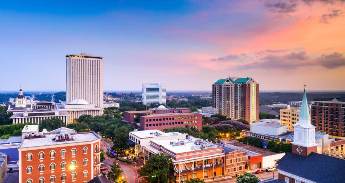 Skyline of a city at sunset featuring a mix of historic and modern buildings, including a tall white building with a rounded roof and a high-rise with a green triangular rooftop. The sky is a blend of pink, orange, and blue hues.