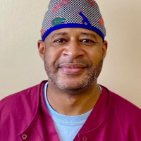 A man wearing a burgundy scrub top and a patterned surgical cap smiles at the camera. He has a neatly trimmed beard and short hair. The background is a plain, light-colored wall.