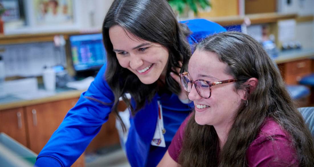 Two women are smiling while looking at a computer screen. One woman with long dark hair in a blue outfit is standing and pointing at the screen, while the other woman with long brown hair and glasses is seated and engaged with the computer.