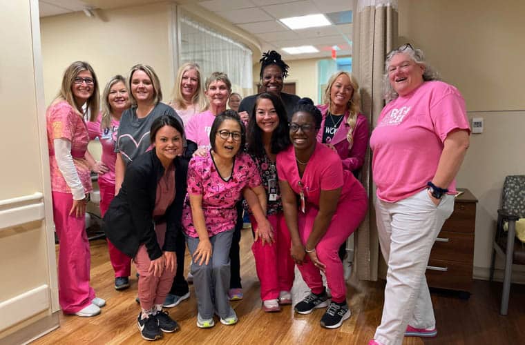 A group of medical staff members posing together in a hospital room. They are all wearing different shades of pink scrubs and casual attire, smiling at the camera. The room has beige walls, curtains, and wooden flooring.