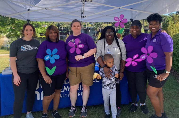 A group of six adults and one child stand under a canopy, posing for a photo. Four adults hold paper flowers, and one of the adults is giving a thumbs-up. They are all smiling and dressed in casual clothes. There is a table with a blue cloth in the background.