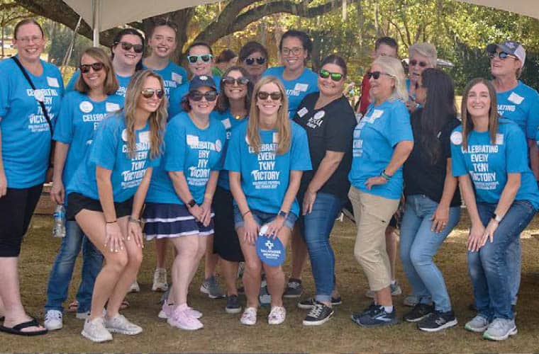 A group of people in blue t-shirts posing and smiling for a photo at an outdoor event. Some of them are wearing sunglasses, and they appear to be under a large white canopy. The t-shirts read “Celebrating Every Tiny Victory.” Trees are visible in the background.