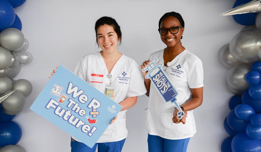 Two healthcare professionals in white uniforms stand between blue and silver balloon columns, smiling and holding signs. One sign reads "We R The Future!" with healthcare-themed icons, and the other is shaped like a large syringe that says, "I gave out 5 shots today!.