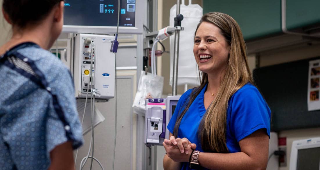 A nurse in blue scrubs smiles and talks to a patient in a hospital room. The patient, wearing a hospital gown, has their back to the camera. Medical equipment and monitors are visible in the background.