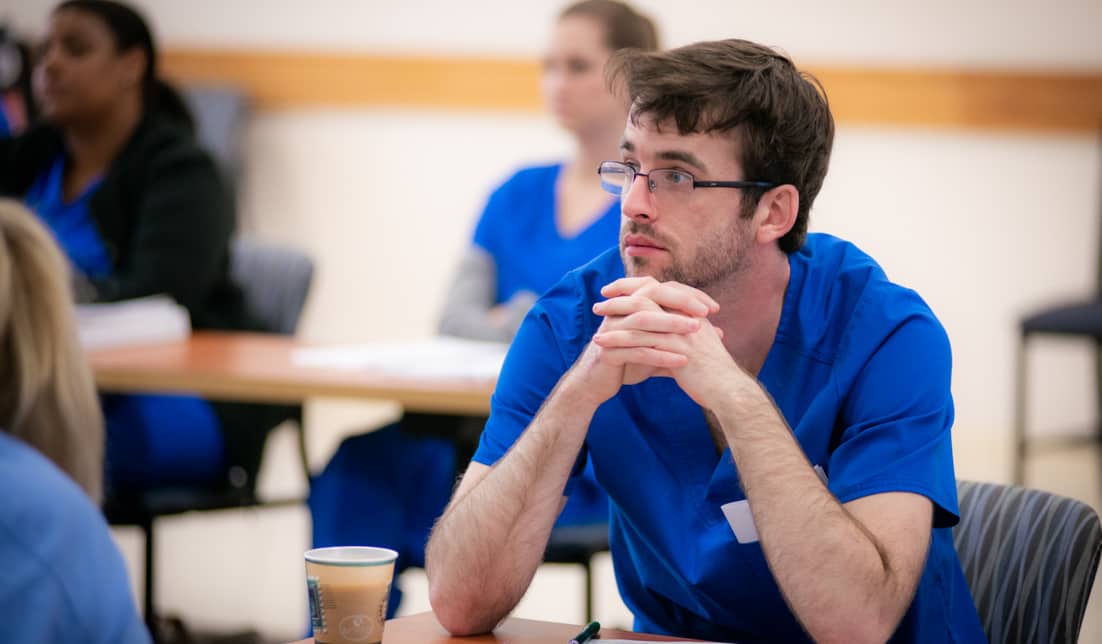 A man in blue medical scrubs sits at a table with his hands clasped together, appearing attentive. Others in similar attire are seated around him, focusing on something out of the frame. A paper cup and papers are on the table in front of him.