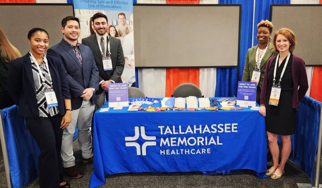 Five professionally dressed individuals stand behind an information booth for Tallahassee Memorial Healthcare at a conference. The booth features brochures, pens, and a large blue tablecloth with the organization's logo. Two large screens are visible in the background.