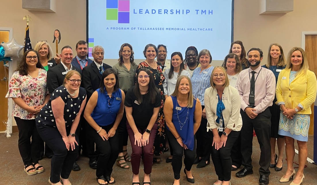 A diverse group of men and women, dressed in business and casual attire, are posing together in front of a screen that reads, "Leadership TMH: A Program of Tallahassee Memorial Healthcare." They are smiling and appear to be in a meeting room.