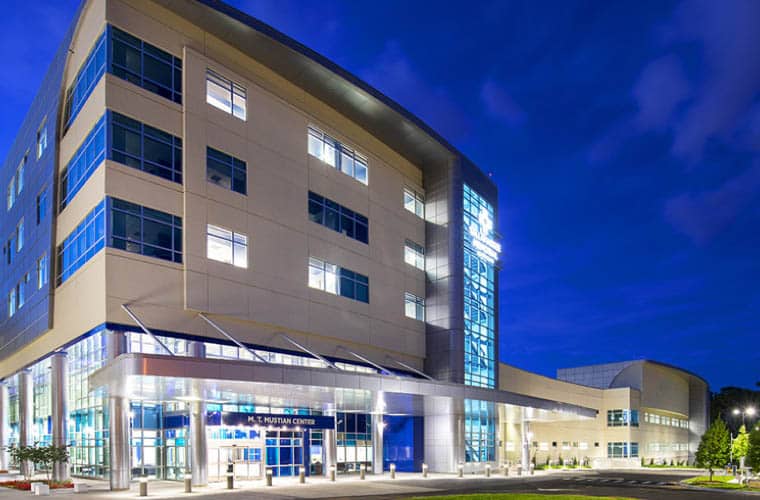 A modern, multistoried building with large windows and a sleek design, illuminated at night. The entrance has a canopy, and the surrounding area is well-lit, enhancing the architectural features against a deep blue evening sky.