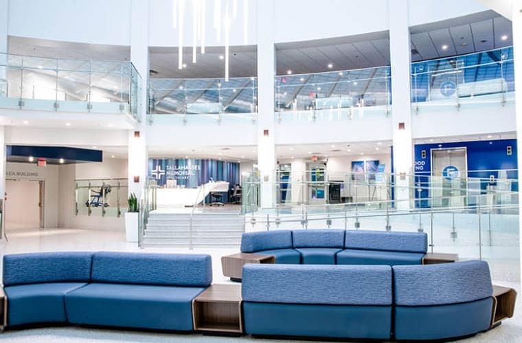 The image shows a modern, spacious lobby area with blue cushioned seating. A central chandelier hangs from the high ceiling. Glass railings line an upper level, and signs indicate various areas including medical services. The area is brightly lit and clean.