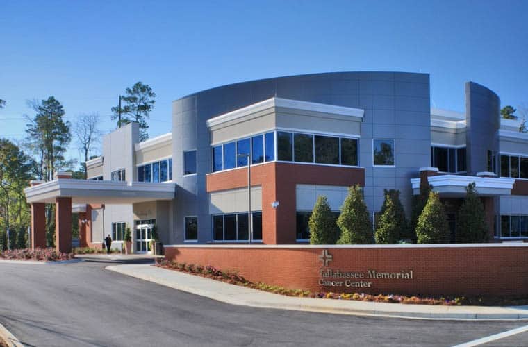 A two-story building with modern architecture features large windows and a brick exterior. The sign reads "Tallahassee Memorial Cancer Center." Surrounding the building are well-maintained shrubs and trees, with a driveway and parking area in front.