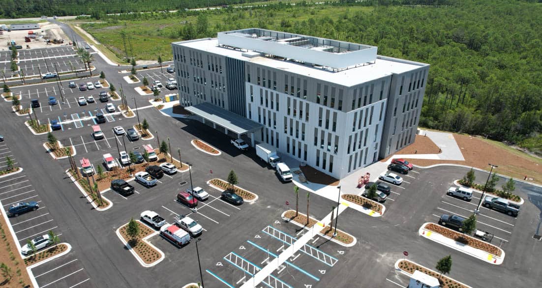 Aerial view of a modern, multi-story building with geometric patterns on its exterior walls and a rooftop structure. The building is surrounded by a large, newly-paved parking lot with numerous cars and landscaped areas. It is set against a backdrop of green trees.