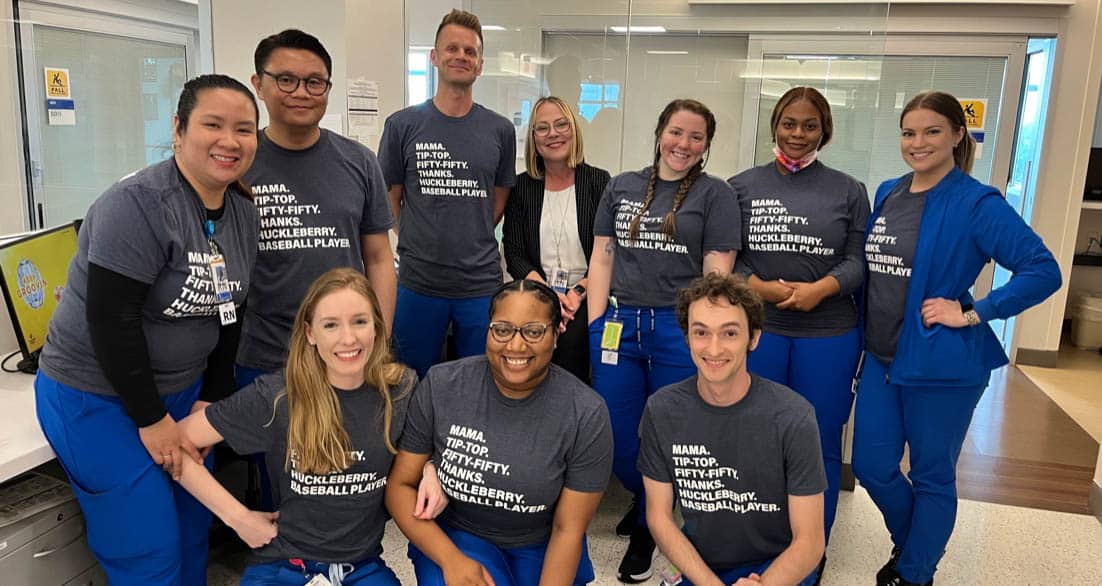 A group of healthcare workers in blue scrubs poses for a photo in a hospital setting. Most are wearing dark gray t-shirts with text on them. They are standing and kneeling, smiling at the camera, with a glass-walled room and hospital equipment in the background.