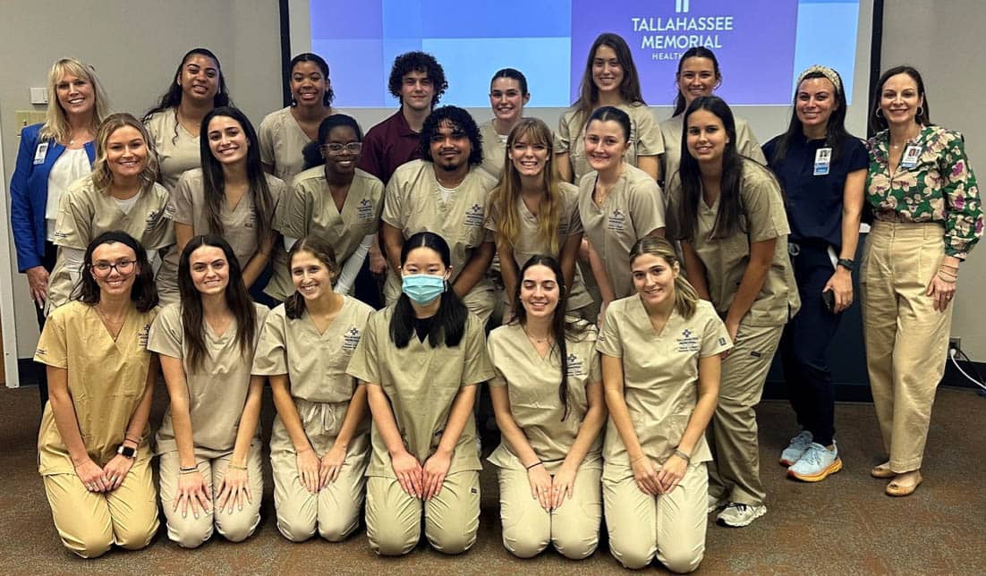 A group of healthcare workers, wearing beige scrubs, poses for a group photo in a conference room. Behind them, a sign reads "Tallahassee Memorial Health." Two people in professional attire and one in blue scrubs stand alongside them.