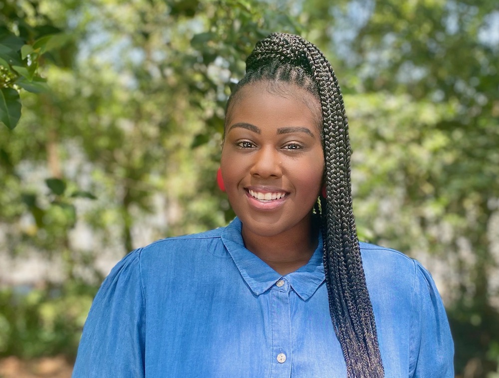 A person with long braids and pink earrings smiles warmly at the camera. They are wearing a blue button-up shirt and standing outdoors with a background of green trees and foliage.