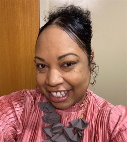 A woman with a cheerful smile takes a selfie. Her curly hair is pulled back, and she wears a pink blouse with a decorative, chunky necklace. The background features a wooden door and a light-colored wall.